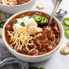 two bowls of chili and cheese soup on a table with crackers, jalapenos, and sour cream