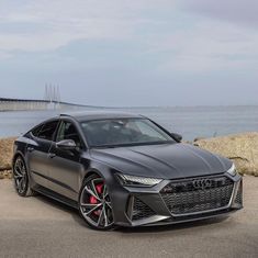 the front end of a gray audi car parked by the water with a bridge in the background