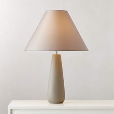 a white table lamp with a beige shade on it's base and an off - white wall in the background