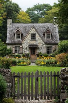 a stone house with a wooden fence and flowers in the front yard, surrounded by greenery