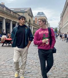 two young men are walking down the street in front of some buildings, one is holding a drink