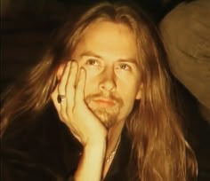 a man with long hair sitting down and looking at the camera while holding his hand to his face