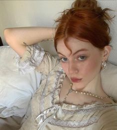 a woman with red hair laying on top of a bed wearing a white dress and pearls