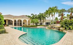 a large swimming pool surrounded by palm trees and shrubs in front of a house with a covered patio