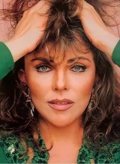 a close up of a person with long hair and blue eyes wearing green shirt holding her hands to her head