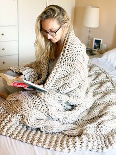 a woman sitting on a bed reading a book while wearing glasses and a knitted blanket