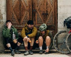 three people sitting on the side of a building next to each other with bikes in front of them