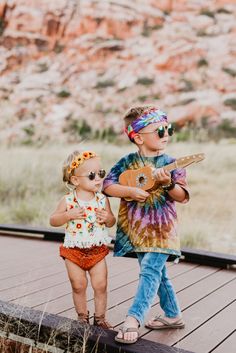 two young children standing on a wooden deck playing guitar and wearing colorful clothing with mountains in the background