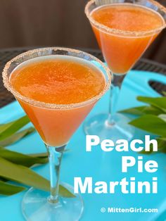 two glasses filled with peach pie martini sitting on top of a blue tray next to green leaves