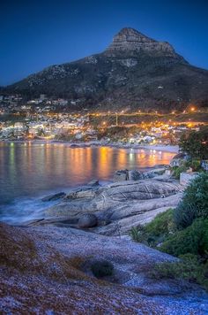 the city lights shine brightly in the night sky over water and rocks, with mountains in the background