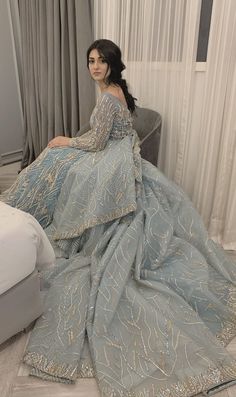 a woman in a blue dress sitting on a bed next to a gray couch and white curtains