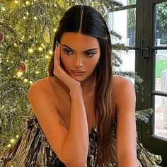 a woman sitting in front of a christmas tree with her hand on her face and looking at the camera