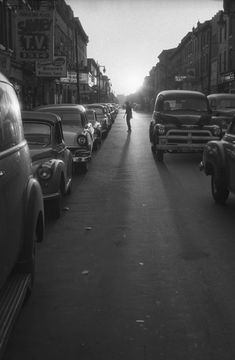 an old black and white photo of cars parked on the side of a street with a person walking down it