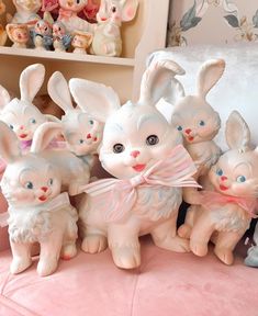 a group of ceramic rabbits sitting next to each other on a pink bed spread with stuffed animals in the background