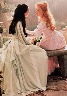 two women in dresses sitting on a bench talking and looking at each other's backs