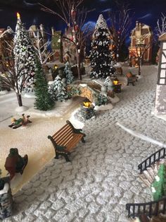 a miniature christmas village with people sitting on benches and snow covered trees in the background