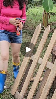 a woman in pink shirt and blue boots standing next to a wooden structure with plants