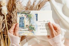 a person holding up a wedding card in front of some dried plants