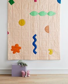 a quilted wall hanging on the side of a white wall next to a teddy bear