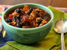 a green bowl filled with chili and beans on top of a table next to a spoon