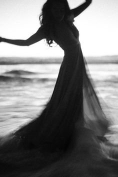 a woman in a long dress is standing on the beach with her arms spread out