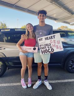 a man and woman standing next to each other in front of a car holding a sign