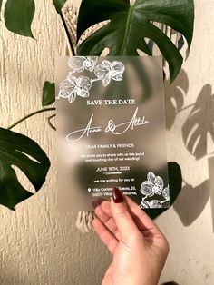 a person holding up a save the date card in front of a potted plant