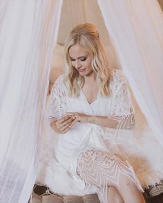 a woman sitting on top of a bed next to a white netted canopy and looking at her cell phone