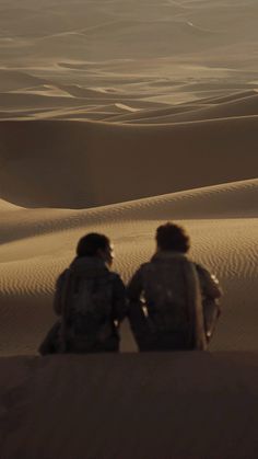 two people sitting on top of a sand dune in the desert, looking at something