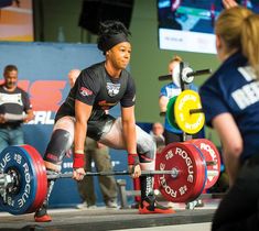 a woman squats while holding a barbell in front of her and other people watching