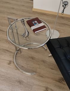 a glass and chrome coffee table with a book on it's side stand next to a black leather couch