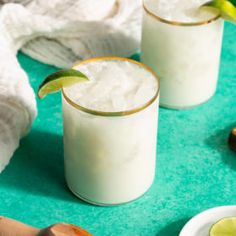 two glasses filled with white liquid and limes on top of a green tablecloth