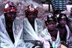 four women wearing head wraps and scarves are sitting on the ground in front of some rocks