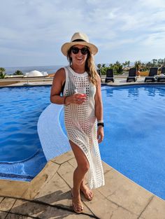 a woman standing in front of a swimming pool wearing a hat and sunglasses, holding a drink