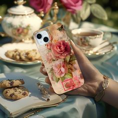 a woman holding up her phone case with flowers on it next to cookies and teacups