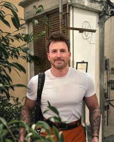 a man standing in front of a building wearing an orange pants and white shirt with tattoos on his arms