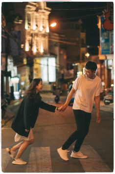 two people holding hands crossing the street in front of a building at night with lights on