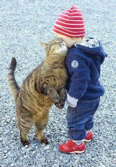 a small child standing next to a cat on top of gravel covered ground and touching it's face