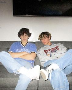 two young men sitting on top of a gray couch in front of a flat screen tv