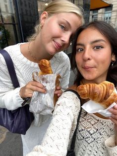 two women are holding croissants and posing for the camera on the street