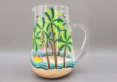 a glass pitcher with palm trees painted on the front and sides, sitting on a gray surface