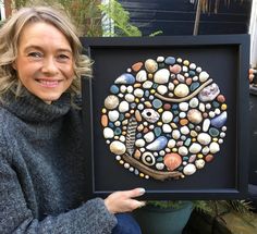 a woman holding up a framed artwork with rocks and shells on it's face