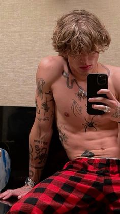 a shirtless man sitting on a couch looking at his cell phone while wearing plaid pants