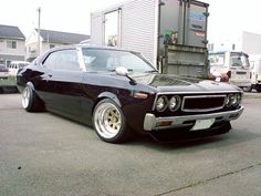 a black muscle car parked in front of a garage with other cars and trucks behind it