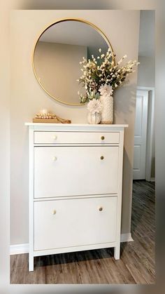 a white dresser with flowers on top and a round mirror above it in a room