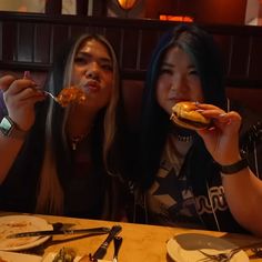 two women sitting at a table with food in their hands and one holding a fork up to her mouth