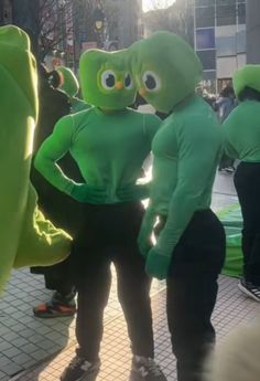 two people in green costumes standing next to each other on a sidewalk with one person wearing an alien costume