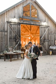 a bride and groom standing in front of a barn with lights on the side of it