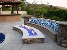 an outdoor fire pit and seating area with blue pillows on the seat wall next to it