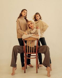 three women in sweaters sitting on a wooden chair with their arms around each other